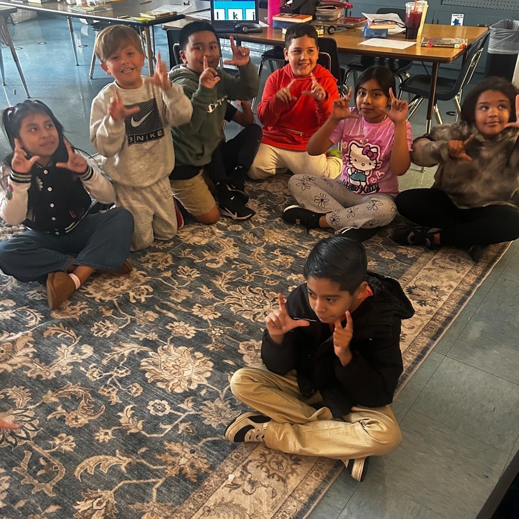 A group of smiling elementary students sit in a circle on a classroom rug, practicing a hand sign together.