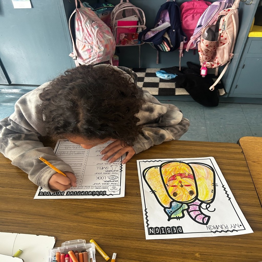 A student sits at a desk coloring a pumpkin drawing and writing descriptive details on a worksheet. Backpacks hang on hooks in the background.