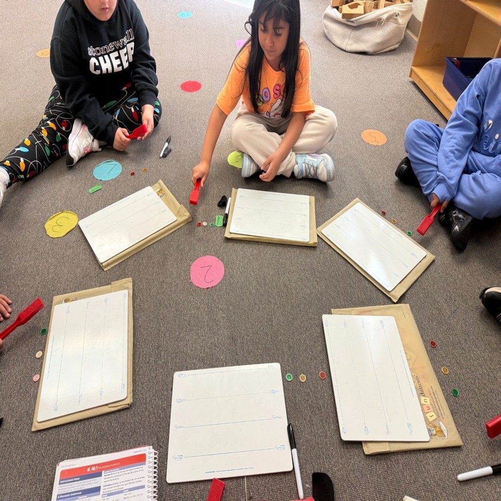 Students sit in a circle on the classroom floor using whiteboards, markers, and small colored chips for a word-building lesson.