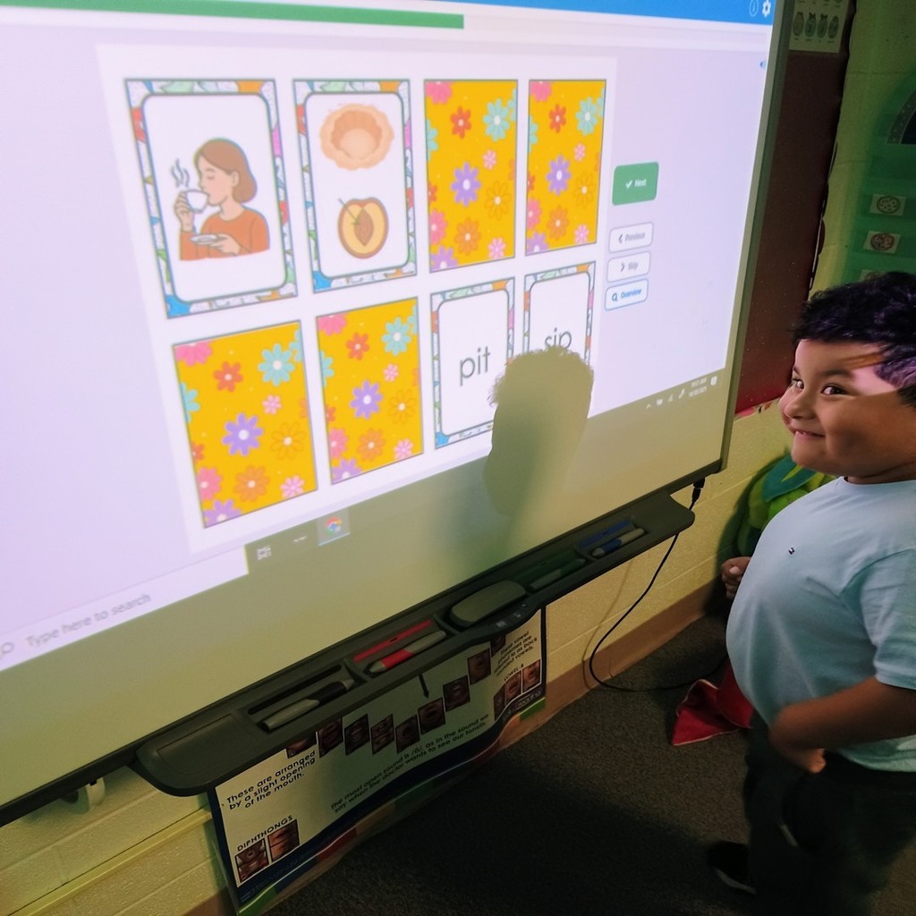 A young student smiles while standing in front of a smartboard showing a word and picture-matching game. The screen displays cards with words like “pit” and “sip,” along with images representing the words. The student’s reflection is visible on the whiteboard.
