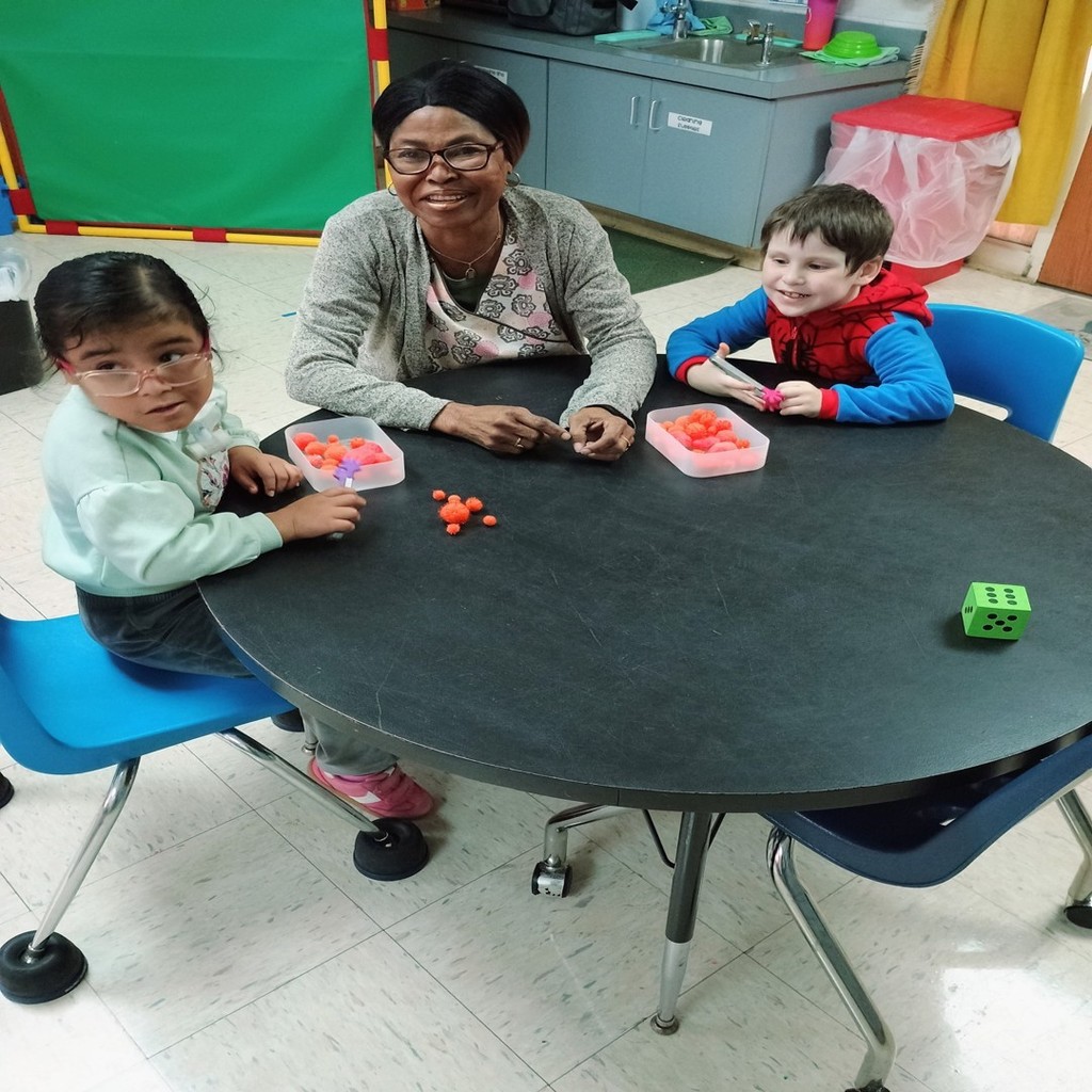 A smiling teacher sits at a round table with two young students during a small-group activity. Each child is holding bright orange pom-poms and sorting them into containers. The classroom background includes a sink, storage cabinets, and colorful learning materials.