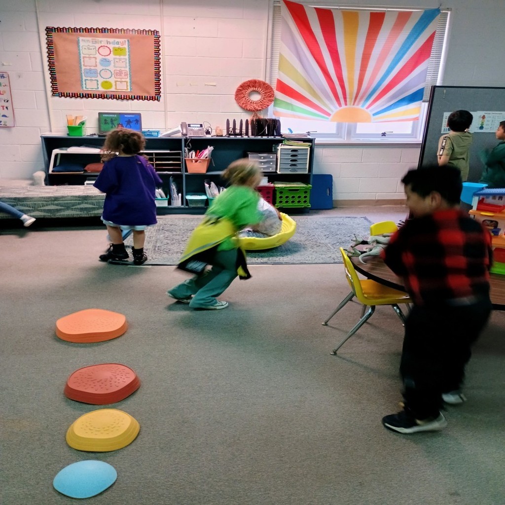 Several children move and play during a classroom activity that includes colorful plastic stepping stones arranged in a line on the carpet. The classroom is decorated with a bright rainbow curtain, posters, and shelves of books and supplies, creating a cheerful environment.