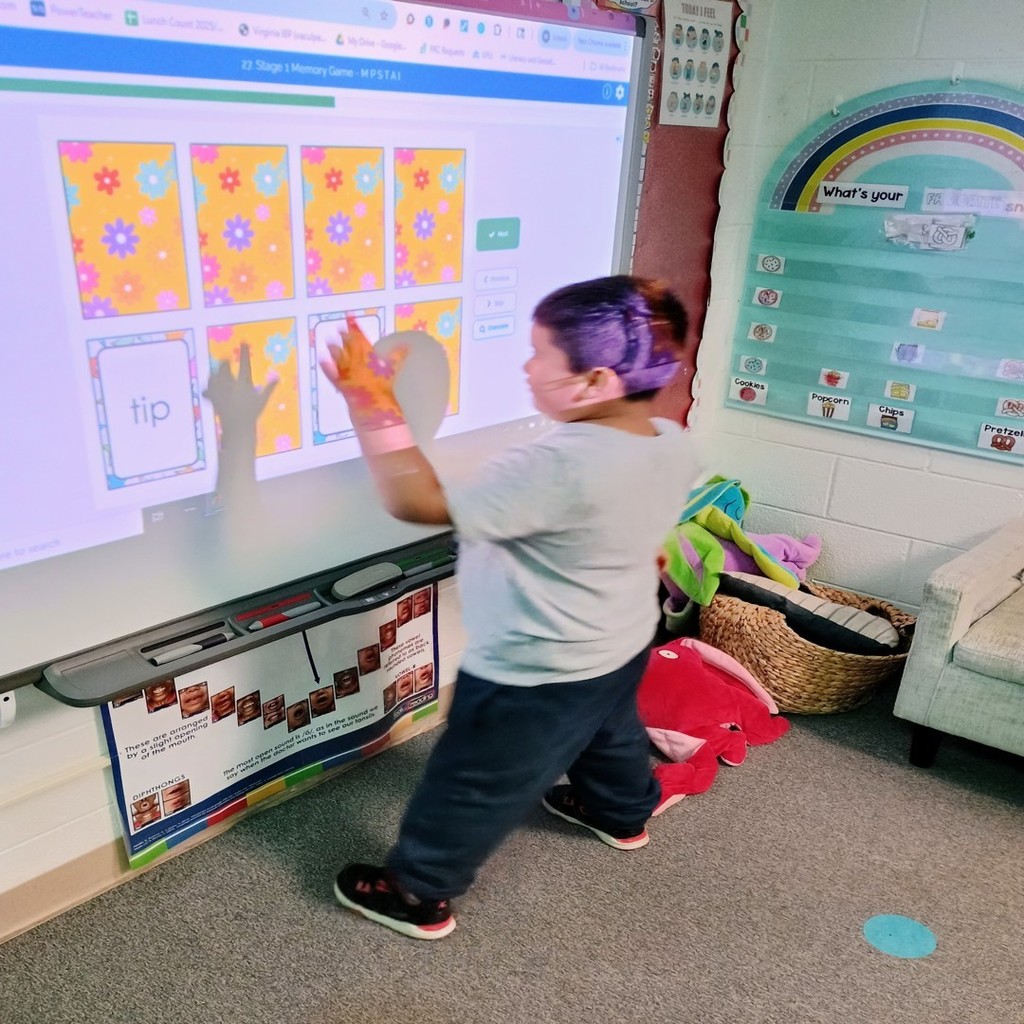A student participates in a digital learning game projected on a smartboard. The screen shows bright yellow and orange flower-patterned cards used for a matching game. The student is reaching out to touch a card while focusing on the activity.