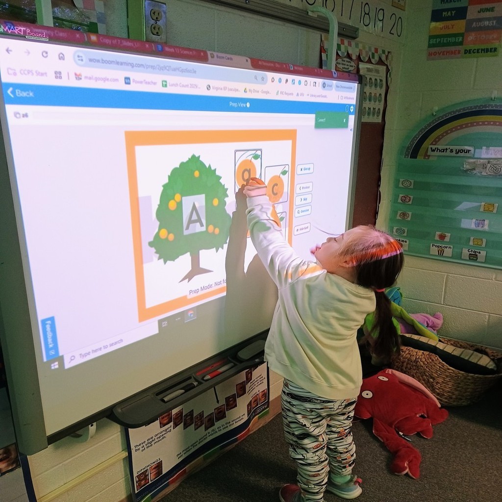 A student stands in front of a classroom smartboard, using a stylus to touch an interactive activity featuring an orange tree and letter cards. The display shows uppercase and lowercase letters, and the student is matching them as part of a literacy lesson.