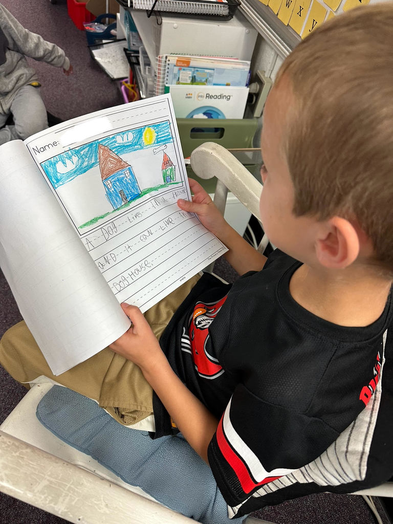 A student wearing a black hoodie sits in a white rocking chair, holding a drawing of a tree and reading from their writing notebook. Letter-sound cards with vowels and digraphs are displayed on the classroom whiteboard.