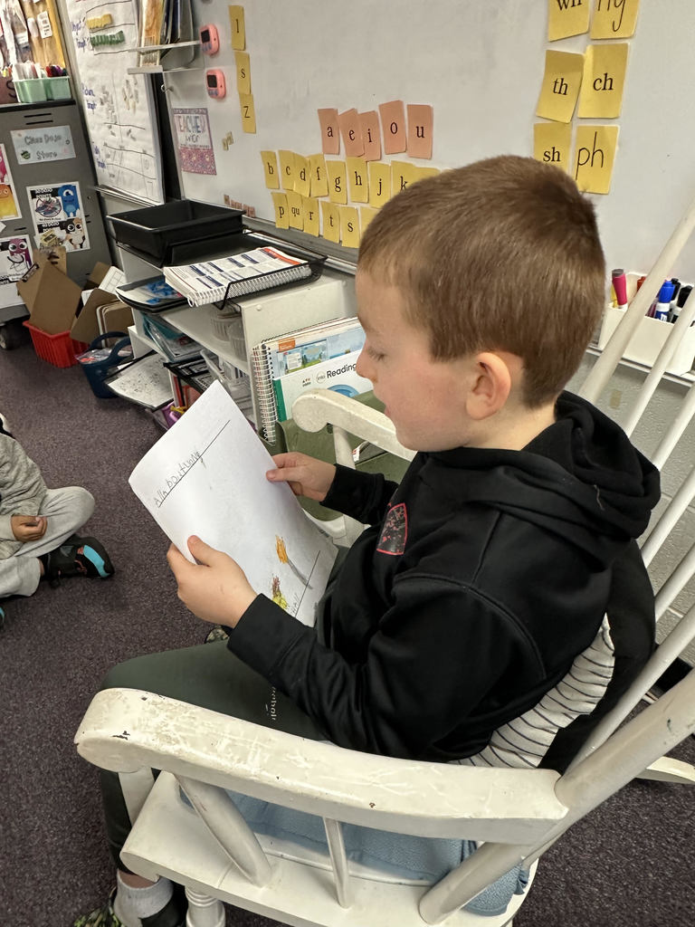 A student in a black shirt sits in a white rocking chair, reading from a handwriting notebook that includes a colorful drawing of two houses under a blue sky with a sun. The classroom background includes a whiteboard and reading materials
