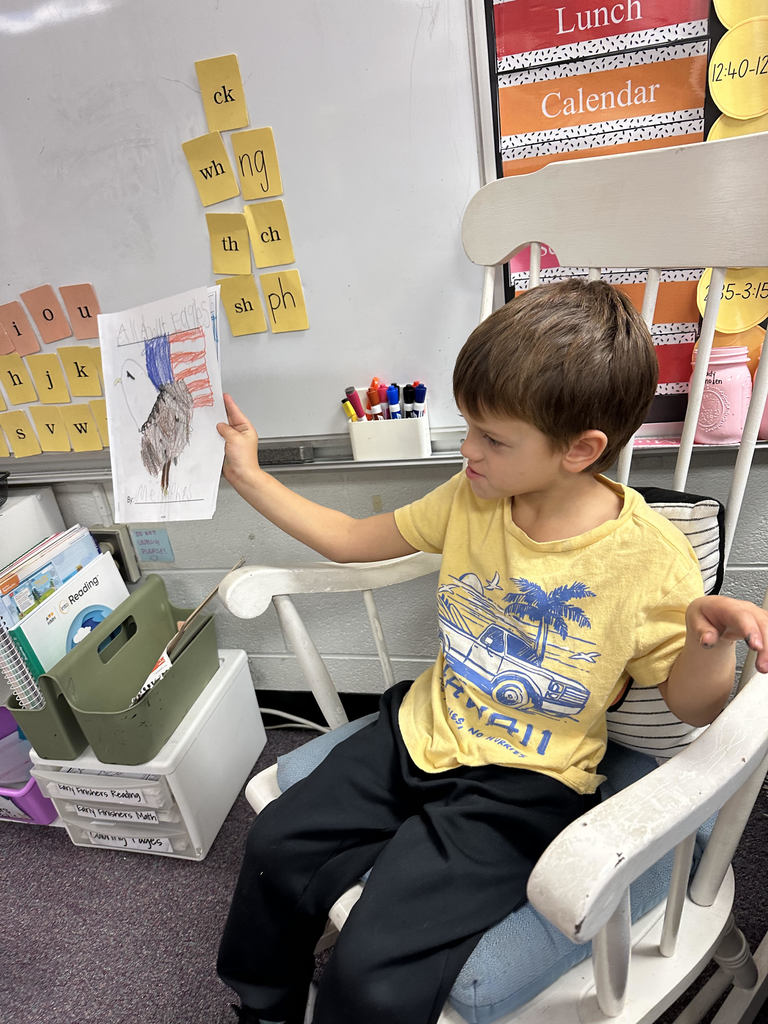 A student wearing a yellow shirt sits in a white rocking chair, reading aloud from a writing notebook. Behind the student is a classroom whiteboard covered with sticky notes displaying letter sounds and word patterns.