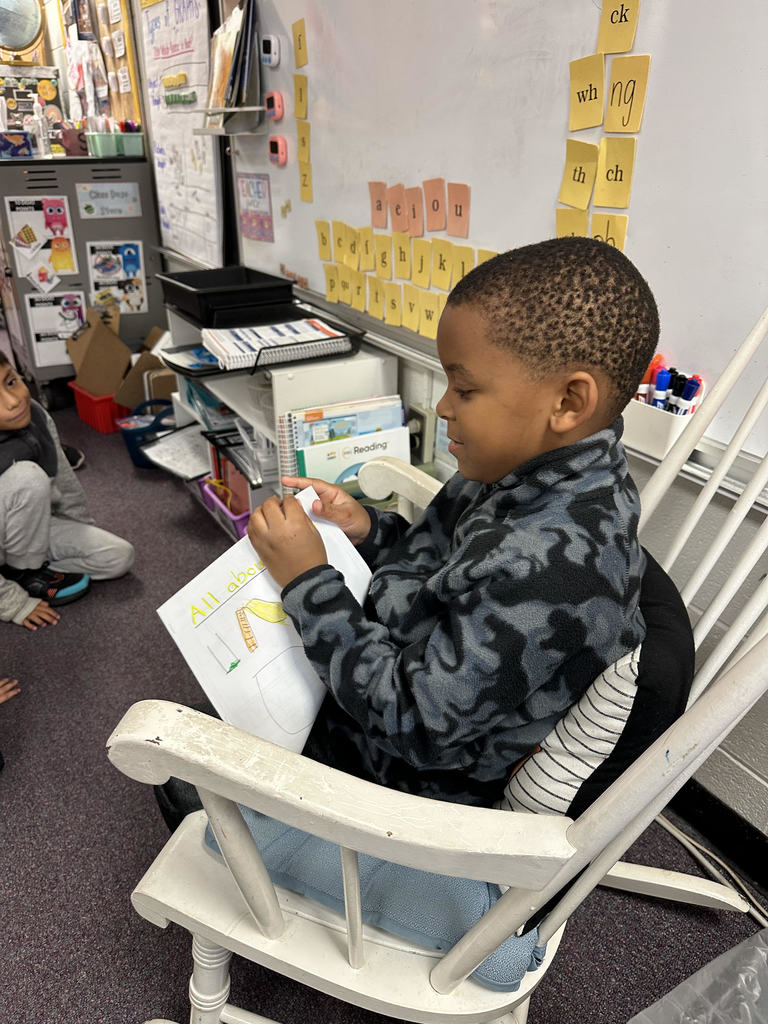 A student in a gray and black patterned sweater reads from a notebook in a white rocking chair. The open page shows a drawing and the title “All About.” Other students are seated nearby, listening to the presentation.