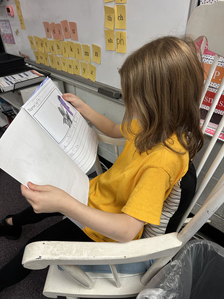 A student in a yellow shirt sits in a white rocking chair, showing a drawing of an eagle in front of an American flag. The page is titled “All About Eagles.” Behind the student, colorful classroom charts and letter-sound cards decorate the whiteboard.