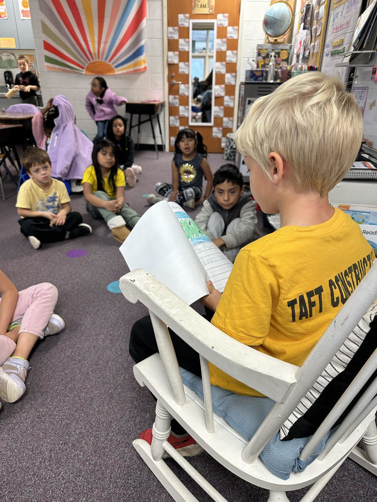 A student in a yellow shirt sits in a white rocking chair at the front of the classroom, reading a story to classmates seated on the carpet. The students watch attentively, and a colorful rainbow curtain hangs near the classroom door.