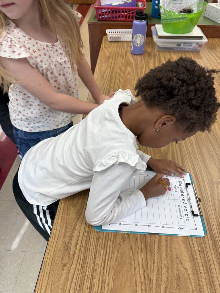 A student leans over a desk to sign a “Registered Voters” sheet on a clipboard while another student waits nearby, watching attentively.