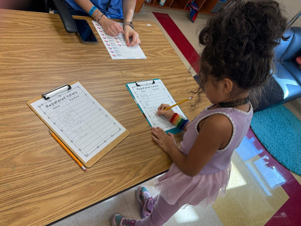 A student dressed in a pink outfit writes her name on a “Registered Voters” sheet as a teacher sits nearby with a sticker sheet ready for new voters.