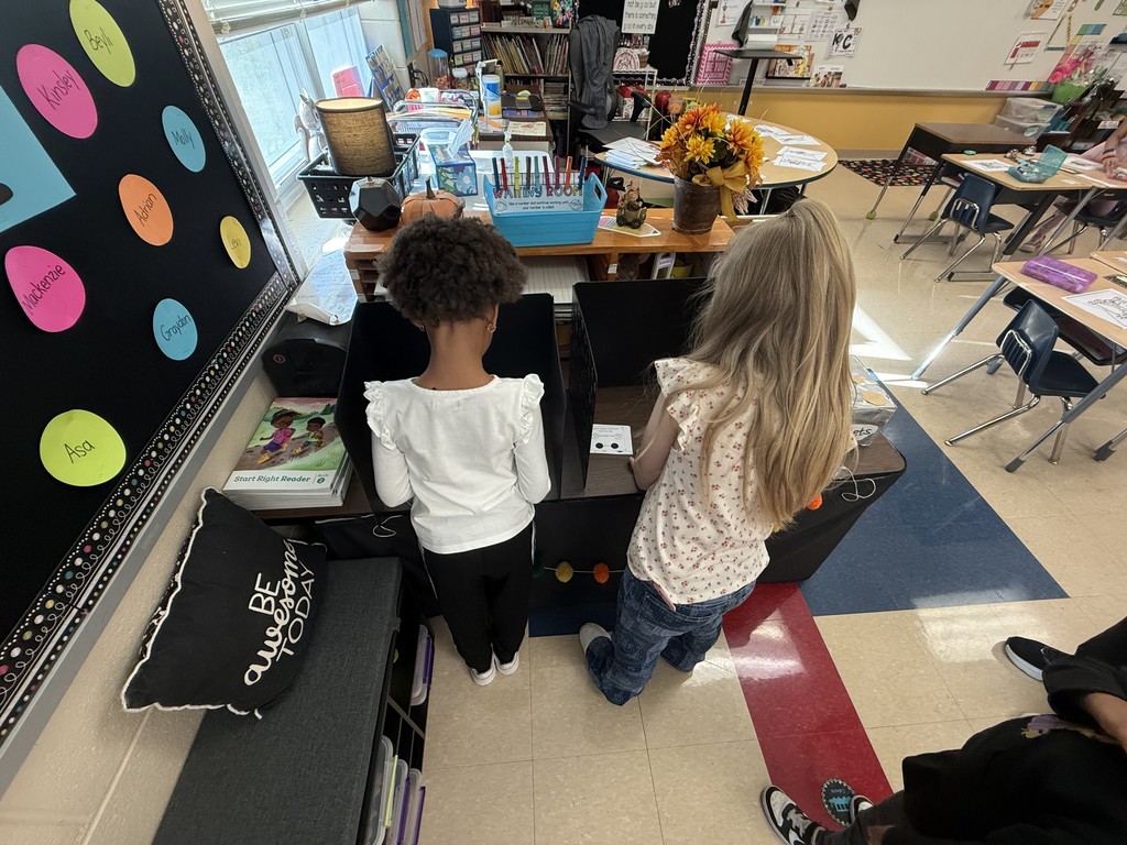 Two students stand side by side at a classroom “voting booth” made from cardboard boxes, casting their ballots in a mock election activity.