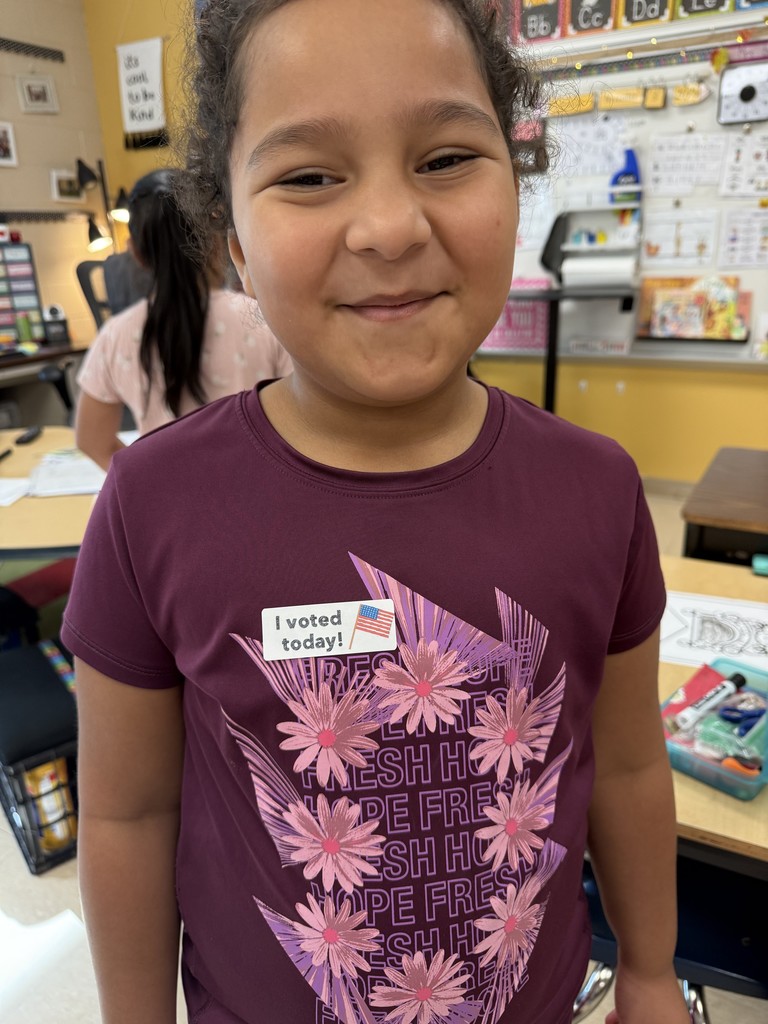 A smiling student wearing a purple shirt decorated with pink flowers proudly shows her “I voted today!” sticker after participating in a classroom voting activity.