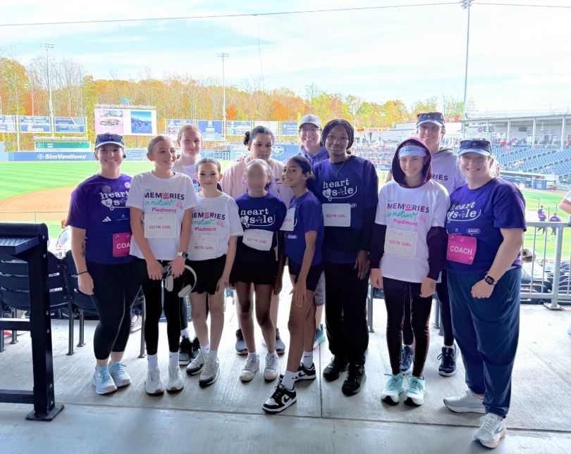 A diverse group of smiling people stands in a stadium, wearing matching athletic gear. Trees with autumn leaves are visible in the background. Energetic and joyful atmosphere.