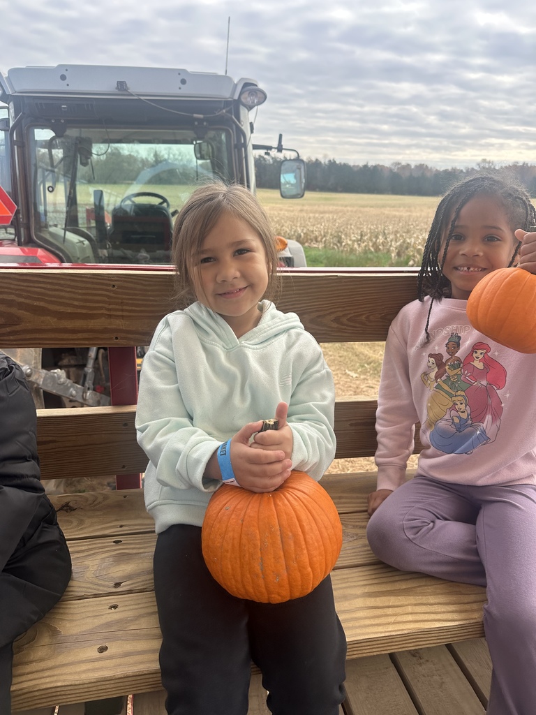 Two smiling children sit side by side on a wooden bench of a wagon, each holding a bright orange pumpkin. A red tractor is parked behind them, and a field of corn stretches out in the background under a cloudy sky.