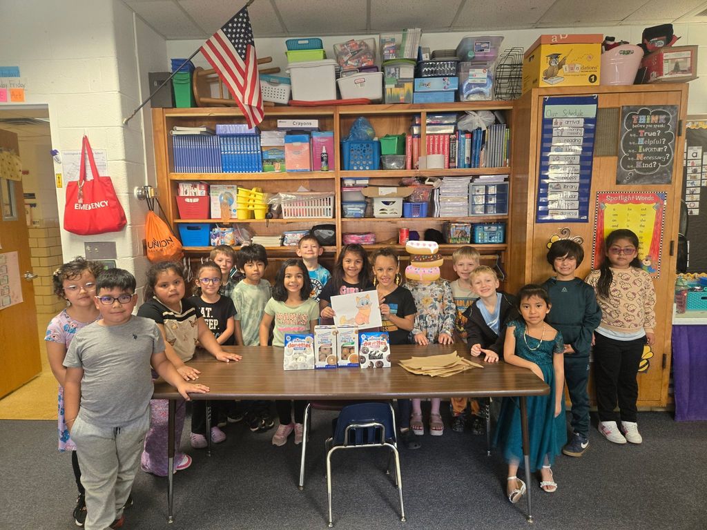 Image contains a class of first graders standing around a table with donuts. Children are smiling. The background contains lots of teacher materials and storage. A red emergency kit bag is hanging on the wall.