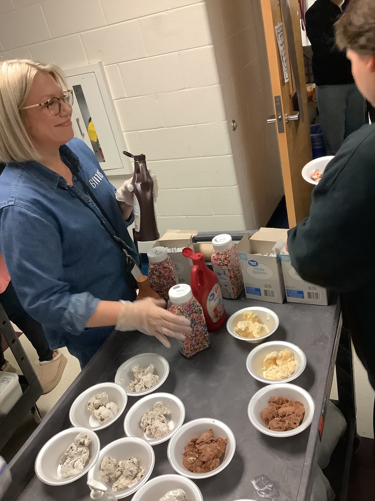 A woman in a blue shirt serves ice cream with toppings like chocolate syrup and sprinkles on a cart. Bowls of chocolate and vanilla ice cream are displayed.
