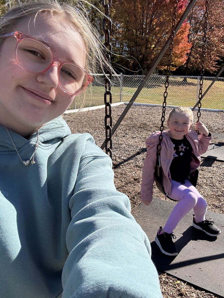 Close-up selfie of a smiling girl with a child sitting on a swing.