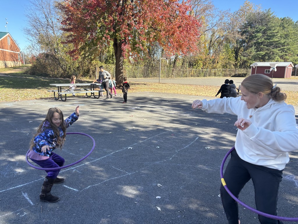 A child in a blue outfit kneeling using a hula hoop with a teacher.