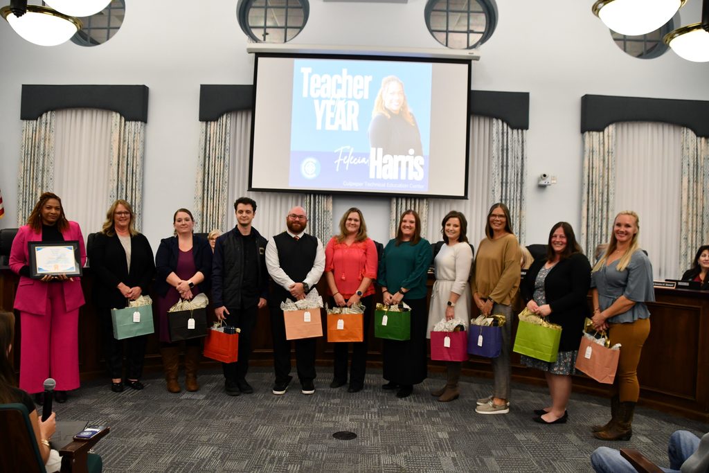 grou p  of teachers standing in a board room holding colorful gift bags with a screen above that says Teacher of the Year