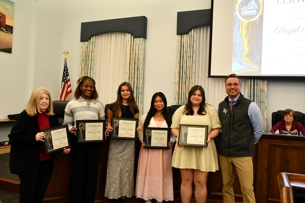 four students standing in a board room holding certificates with a board member on the left and their principal on the right
