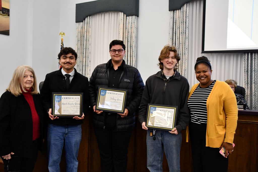 three students holding certificates in board room with board member and teacher