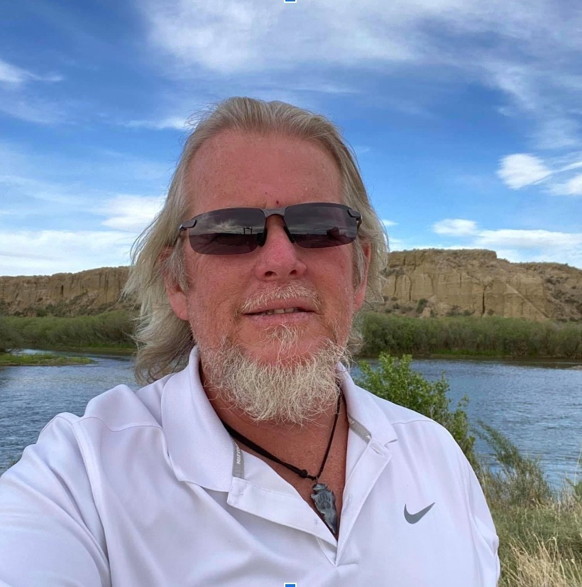 Man with sunglasses and a white shirt stands in front of a serene river, with sandy cliffs and a bright blue sky in the background.
