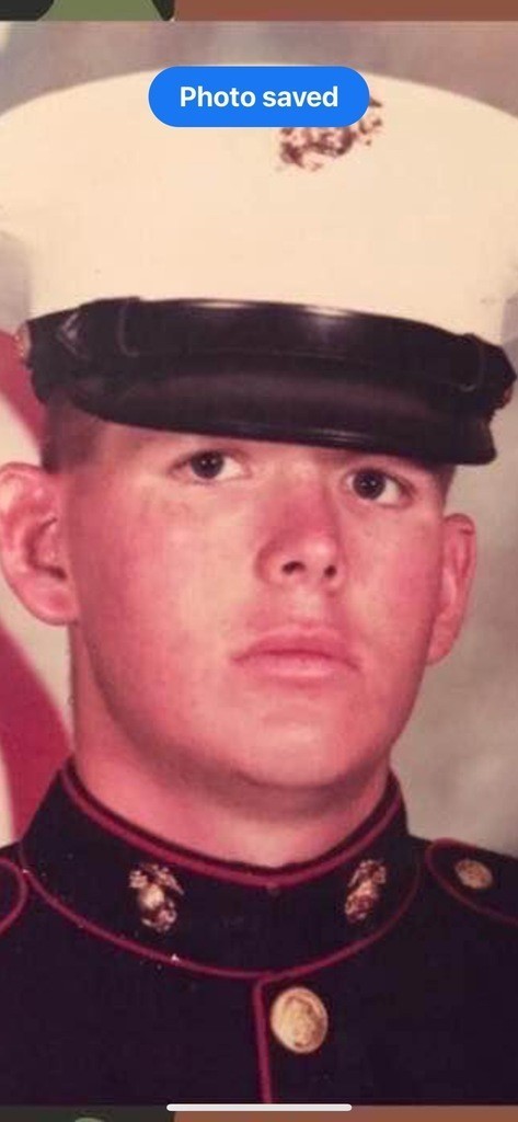 Young man in Marine uniform, wearing a white and black cap. His expression is serious, with a neutral backdrop indicating a formal setting.