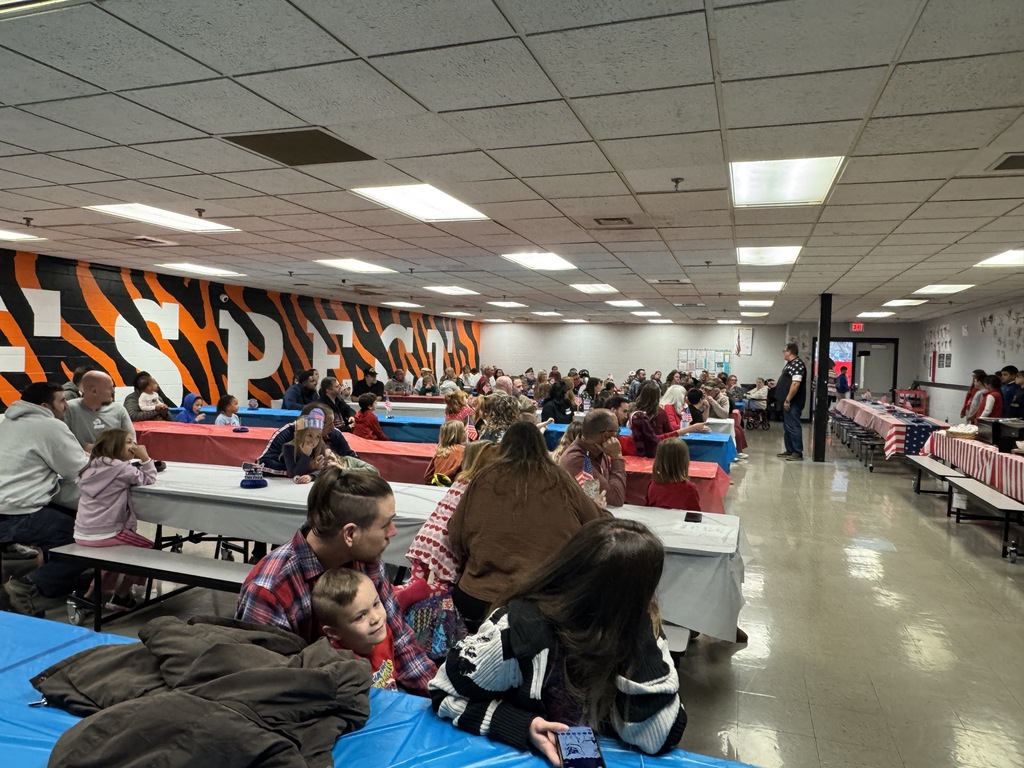 A large group of families and students sitting at tables with blue and red coverings in a cafeteria or large room. An orange and black tiger-stripe mural covers the back wall.