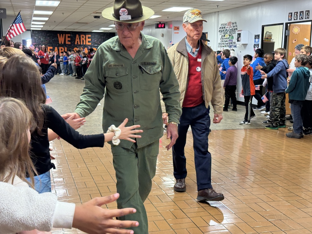 An elderly male veteran in a tan jacket and a VFW cap walking down a school hallway, extending his hand. A large group of excited children, many holding small American flags, line the walls to greet him.