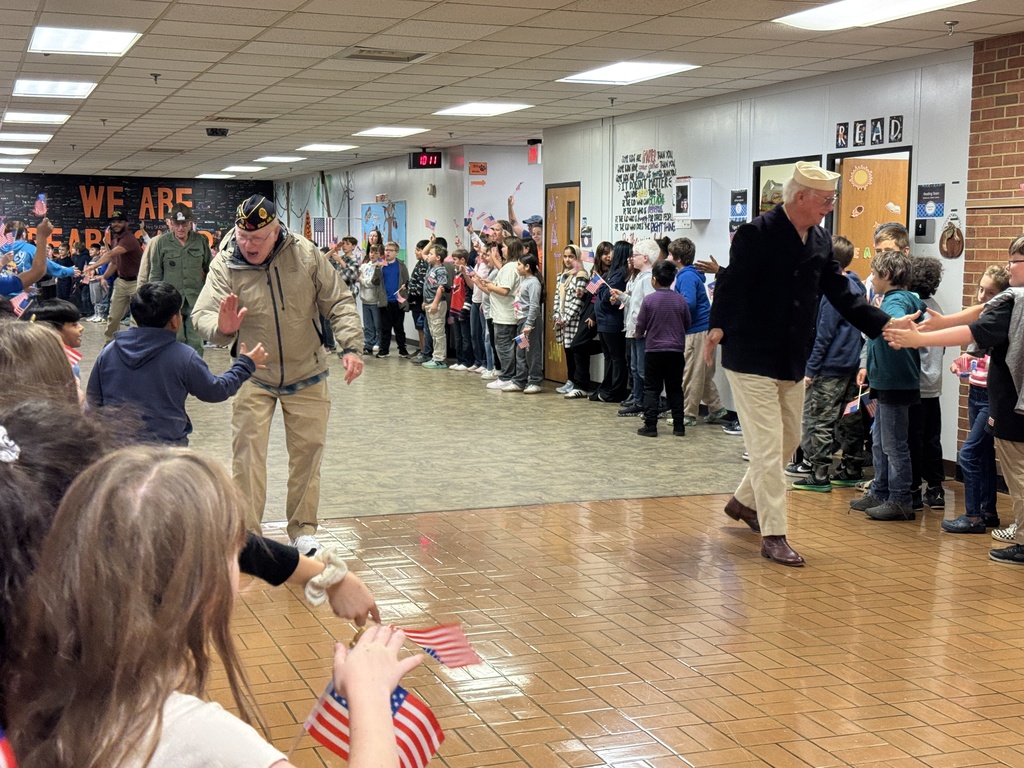 Two male veterans, one in a green military uniform and campaign hat and one in a jacket, walking through a school hallway. Children line the walls reaching out to shake hands.