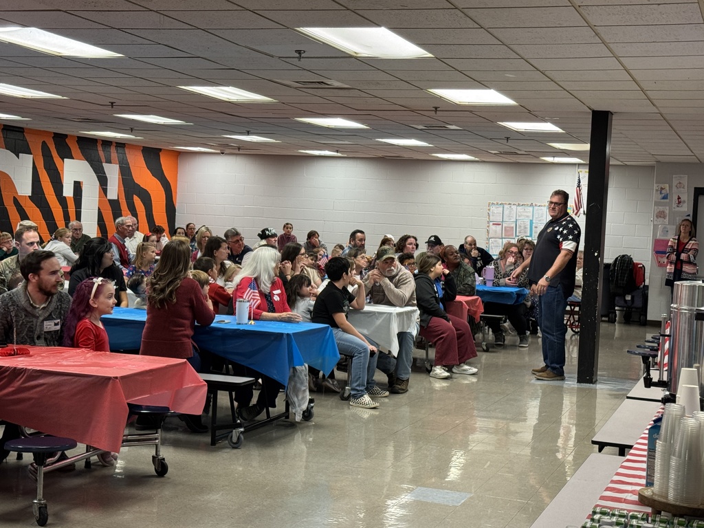 A crowd of people, including families and veterans, seated at tables in a brightly lit cafeteria. A man in a dark shirt is standing near the front addressing the attendees.