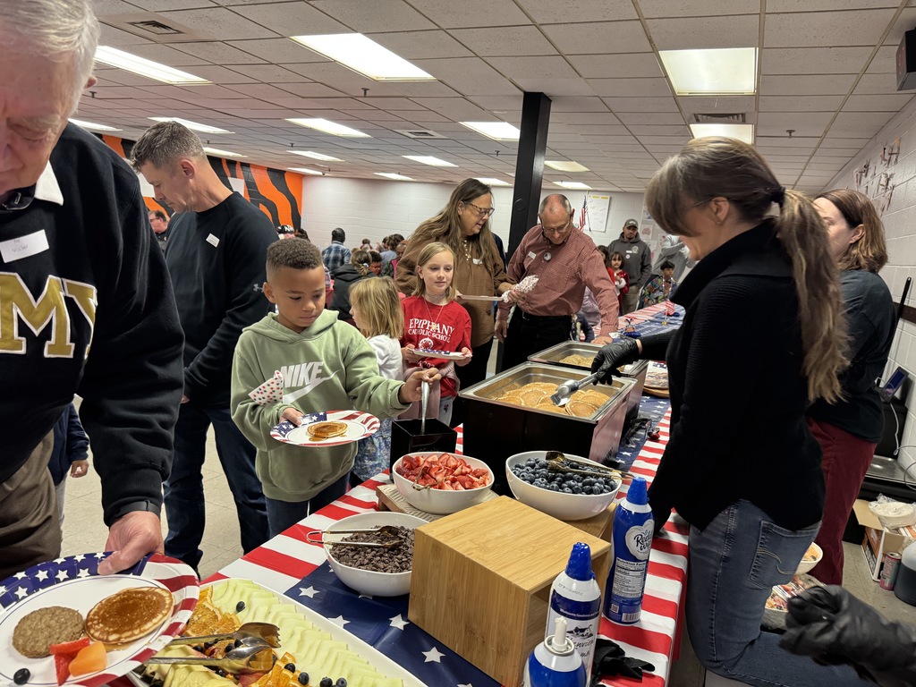 A breakfast buffet line with pancakes, sausage, fruit, and whipped cream. Several people, including children and an elderly man in an "ARMY" sweater, are serving themselves.