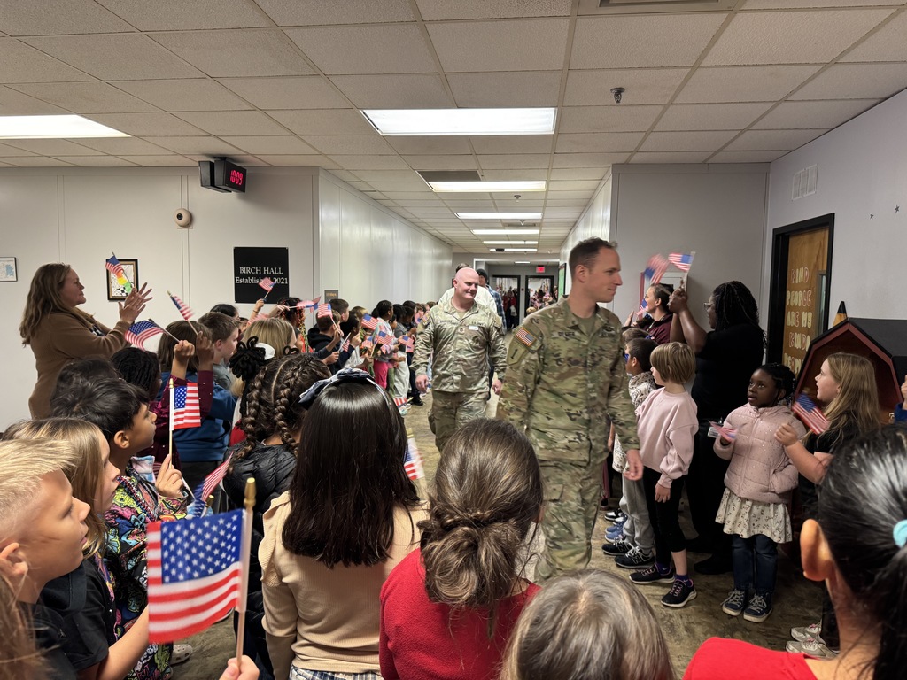 Two male service members in camouflage uniforms walking through a school hallway lined with cheering students waving American flags.