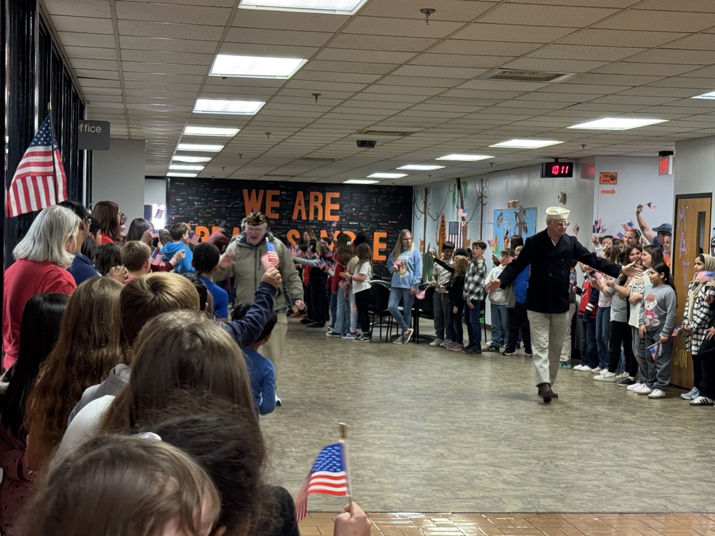 A wide shot of a school hallway where students are lining both sides, waving American flags. Two veterans are walking down the center aisle, receiving applause and greetings. A large sign in the background reads, "WE ARE [...]."