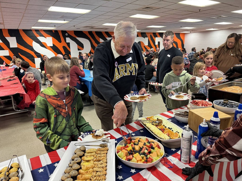 An elderly male veteran in an "ARMY" sweater serving himself from a large table of food, including fruit, pancakes, and pastries. Two young boys are standing next to him, holding plates.