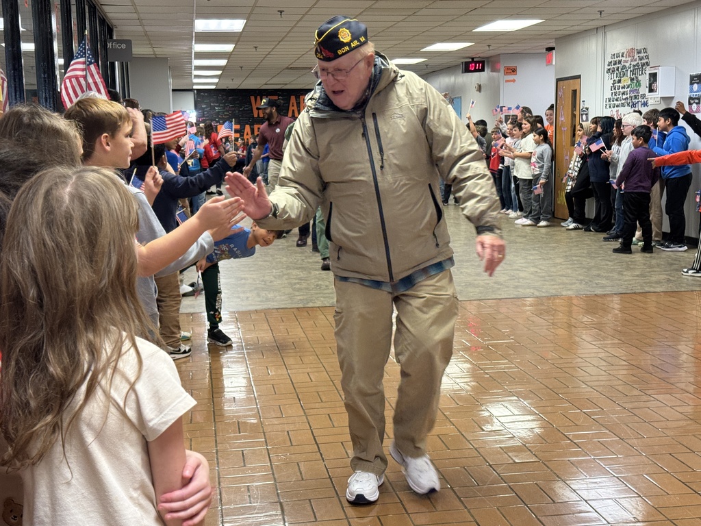 An elderly male veteran in a tan jacket and a VFW cap walking down a school hallway, extending his hand. A large group of excited children, many holding small American flags, line the walls to greet him.