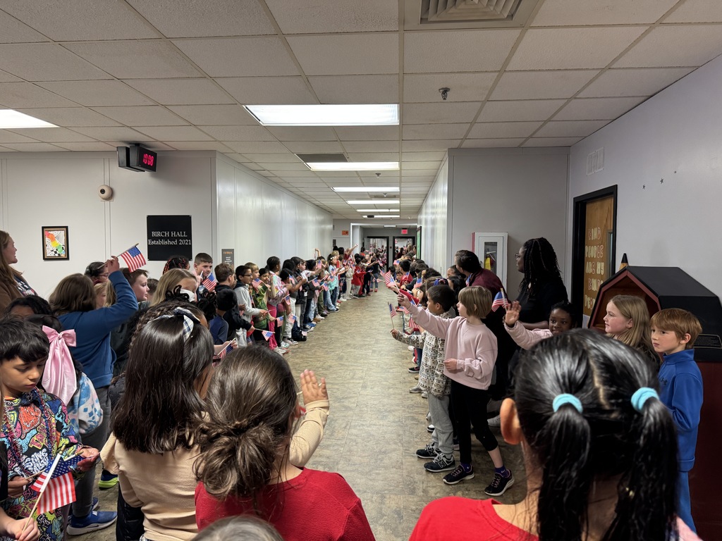 A long view of a school hallway lined on both sides with students facing the center, clapping and waving small American flags. A sign for "BIRCH HALL" is visible on the left.