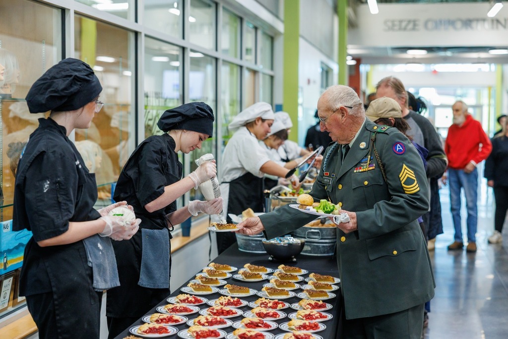 A veteran dressed in uniform receives food from culinary students during the Veterans Day Celebration at the Culpeper Technical Education Center. The students, wearing chef coats and hats, serve an array of plated desserts and meals as part of a luncheon honoring local veterans.