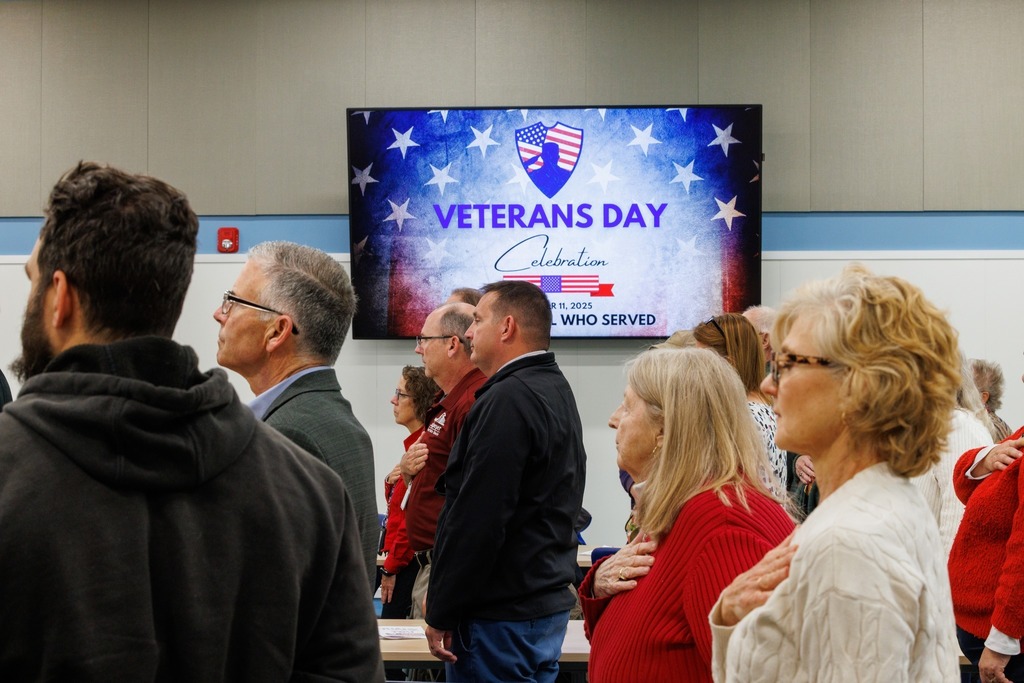Attendees stand with their hands over their hearts during the Veterans Day Celebration at the Culpeper Technical Education Center. A large screen in the background displays the event’s title, “Veterans Day Celebration — Honoring All Who Served,” with an American flag–themed design.