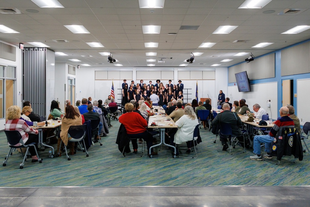 Veterans and guests sit at tables watching a student choir perform on stage during the Veterans Day Celebration at the Culpeper Technical Education Center. The choir, dressed in formal black attire, sings under the direction of their conductor, with American flags displayed on either side of the stage.