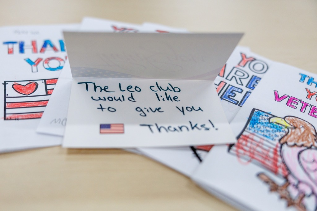 A close-up of colorful handmade thank-you cards created by students for veterans. One open card reads, “The Leo Club would like to give you thanks!” with a small American flag sticker, while others feature hand-colored patriotic designs with messages like “Thank you, veterans.”