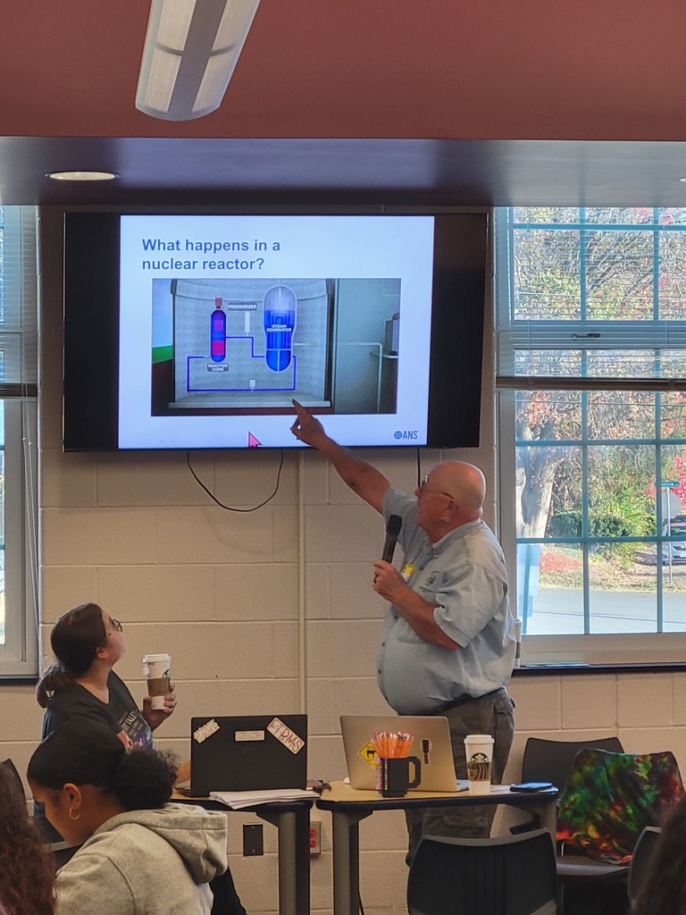 A man presents to a classroom, pointing at a diagram on a screen titled "What happens in a nuclear reactor?" Students sit at desks, watching attentively.