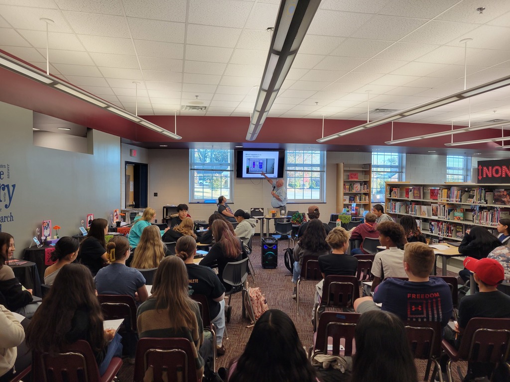 A group of students sits in a library facing a screen, listening to a speaker at the front. Bookshelves and windows in the background create a studious atmosphere.