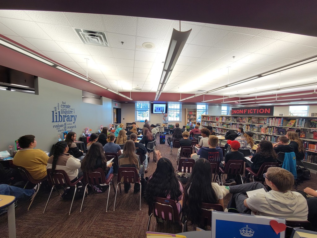 A group of students sits attentively in a bright library, facing a speaker. Bookshelves labeled "Nonfiction" line the walls, creating an academic atmosphere.
