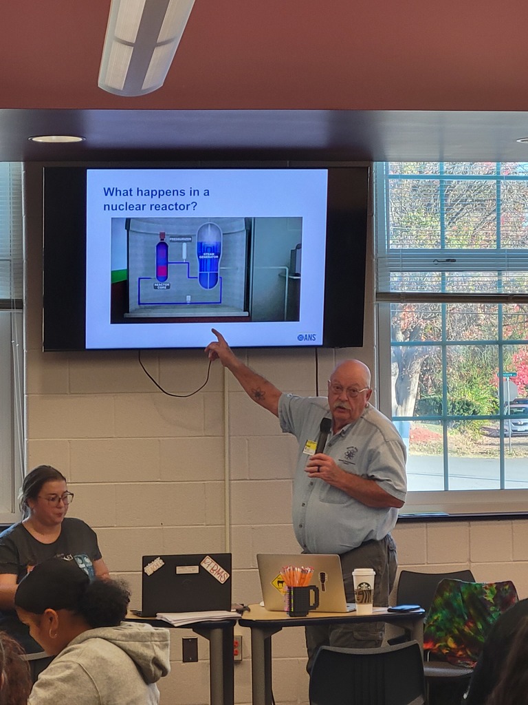 A speaker in a classroom points at a screen displaying a diagram titled "What happens in a nuclear reactor?" as attendees watch attentively.