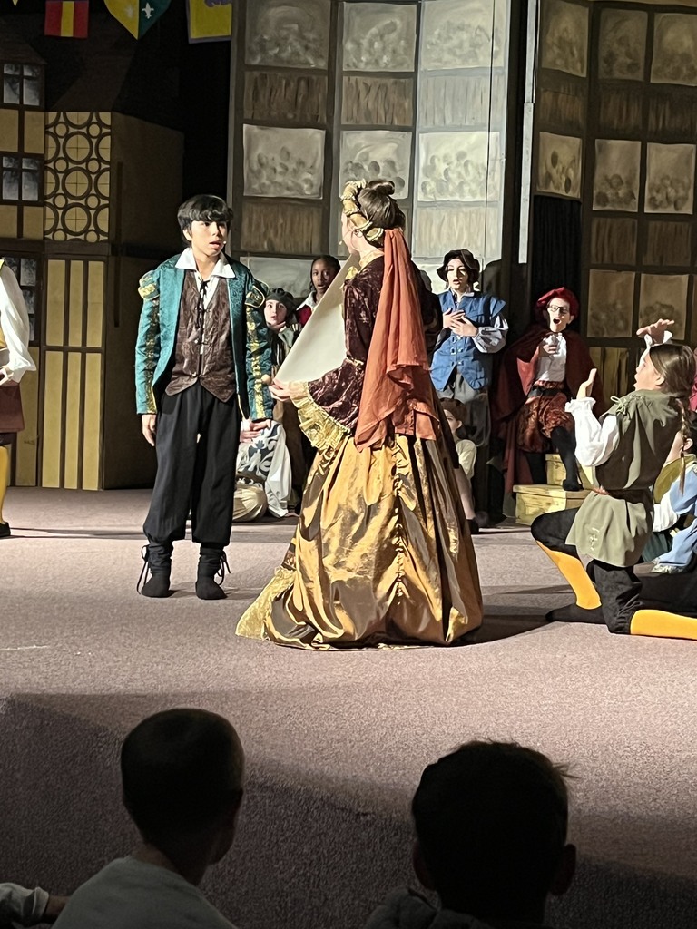 Students perform on stage in period costumes for a school play. A student in a teal jacket and brown vest speaks to another dressed in a gold and rust-colored gown, while other cast members watch in the background. The set features medieval-style painted walls and banners.