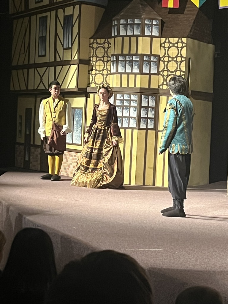 Three student actors stand on stage in front of a painted backdrop resembling a Tudor-style building. One wears yellow knee socks and a brown vest, another wears a gold gown with maroon sleeves, and the third wears a teal jacket. The scene appears to be part of a historical or Shakespearean play.
