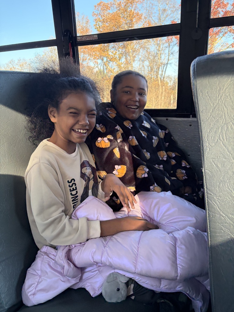Two smiling girls sit together on a school bus seat. They’re wrapped in jackets and blankets, laughing with sunlight streaming through the window. The autumn trees outside glow with orange leaves.